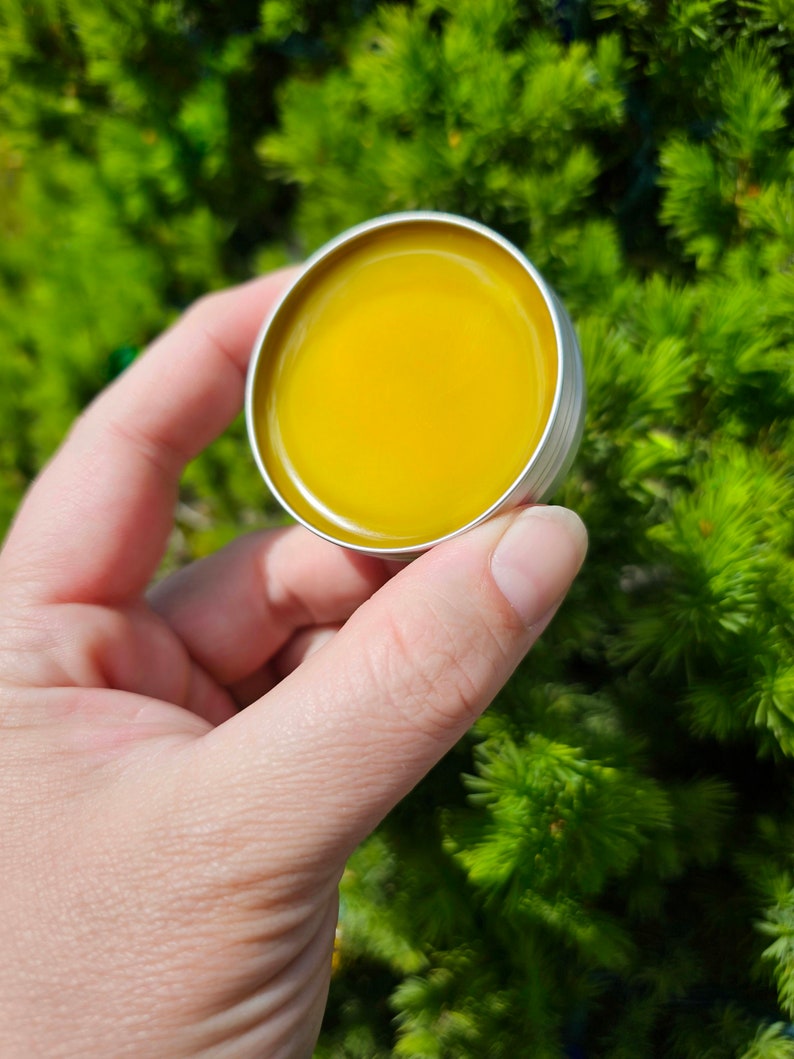 May include: A small round tin container with a yellow balm inside. The tin is held in a hand against a green background.