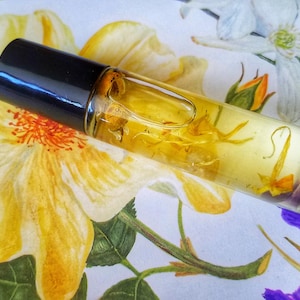 Puede incluir: Un frasco de vidrio transparente con rodillo y tapa negra, lleno de un líquido amarillo y pequeños pétalos de flores blancas. El frasco está sobre un fondo blanco con un patrón floral.