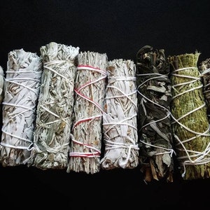 May include: A collection of eight white sage smudge sticks, some tied with white string and others with red string. The smudge sticks are arranged in a row on a black background.