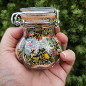 May include: A small, clear glass jar filled with dried herbs and flowers, held in a hand. The jar has a metal clasp and an orange rubber seal. The contents include pink rose petals, blue lavender, and yellow chamomile. The background is a blurred green.