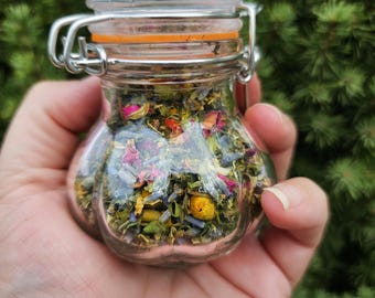 May include: A small, clear glass jar filled with dried herbs and flowers, held in a hand. The jar has a metal clasp and an orange rubber seal. The contents include pink rose petals, blue lavender, and yellow chamomile. The background is a blurred green.