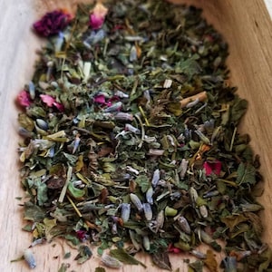 May include: A close-up of a wooden scoop filled with a blend of dried herbs and flowers. The blend includes green herbs, lavender buds, and pink rose petals.