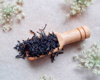 May include: A wooden scoop filled with loose leaf black tea on a light brown surface with white flowers scattered around it.