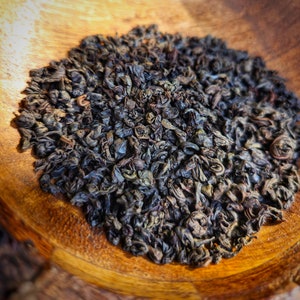 May include: Close-up of a pile of loose leaf black tea on a wooden surface. The tea leaves are dark brown and have a slightly curled shape.