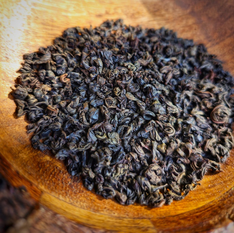 May include: Close-up of a pile of loose leaf black tea on a wooden surface. The tea leaves are dark brown and have a slightly curled shape.