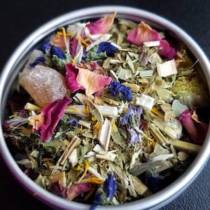 Puede incluir: Una lata redonda plateada llena de una colorida mezcla de hierbas y flores secas. La mezcla incluye pétalos de rosa rosas, flores azules y varias hojas y tallos verdes, lo que sugiere una mezcla de té de hierbas.