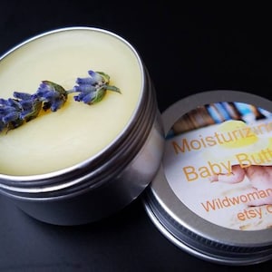 May include: A small silver tin with a white label that reads "Moisturizing Baby Butter" with a picture of a baby on the beach. The tin is open and filled with a yellow cream with a lavender flower on top.