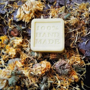 May include: A square bar of soap with the words "100% HAND MADE" printed on it. The soap is surrounded by dried yellow flowers.