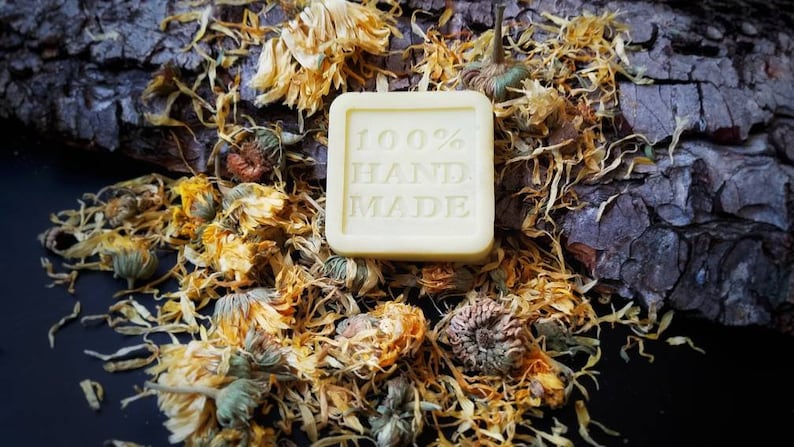 May include: A square bar of soap with the words "100% HAND MADE" printed on it. The soap is surrounded by dried yellow flowers.