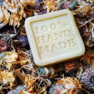 May include: A square, pale yellow bar of soap with the words "100% HAND MADE" embossed on the front. The soap is surrounded by dried yellow flower petals.