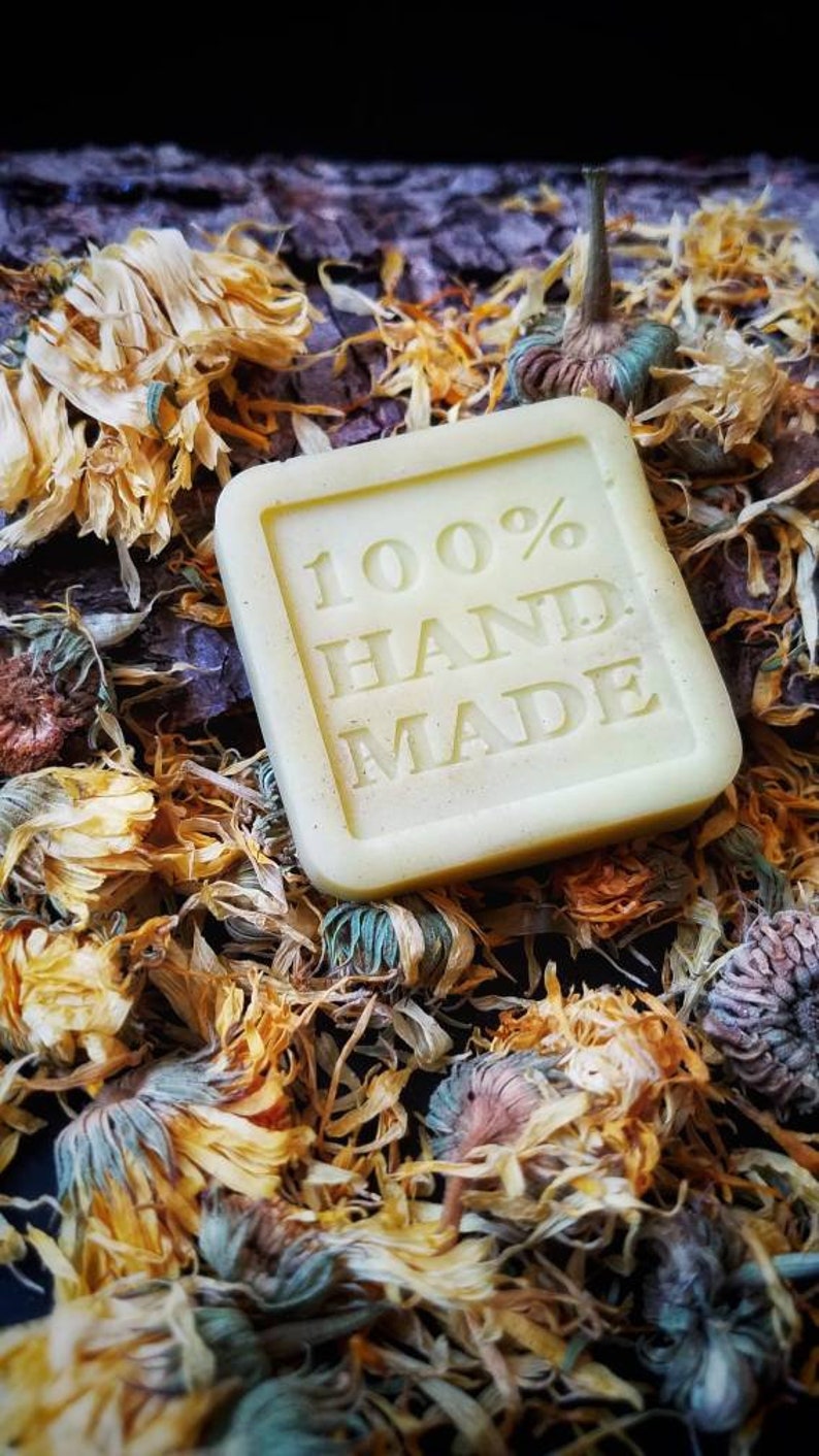 May include: A square, pale yellow bar of soap with the words "100% HAND MADE" embossed on the front. The soap is surrounded by dried yellow flower petals.