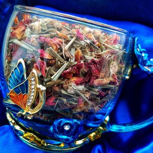 Pode incluir: Uma ch&aacute;vena de ch&aacute; de vidro azul com guarni&ccedil;&atilde;o dourada e um design de borboleta. A ch&aacute;vena est&aacute; cheia de ervas secas e p&eacute;talas de rosa.