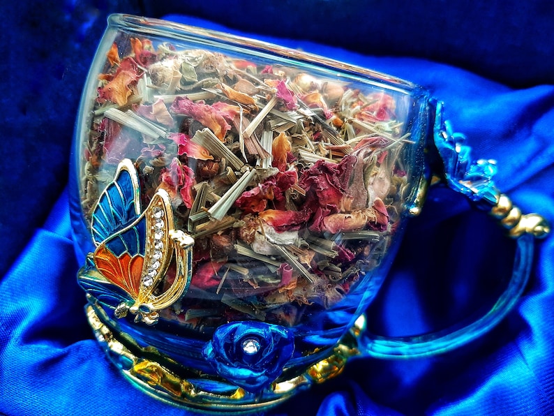 Pode incluir: Uma ch&aacute;vena de ch&aacute; de vidro azul com guarni&ccedil;&atilde;o dourada e um design de borboleta. A ch&aacute;vena est&aacute; cheia de ervas secas e p&eacute;talas de rosa.