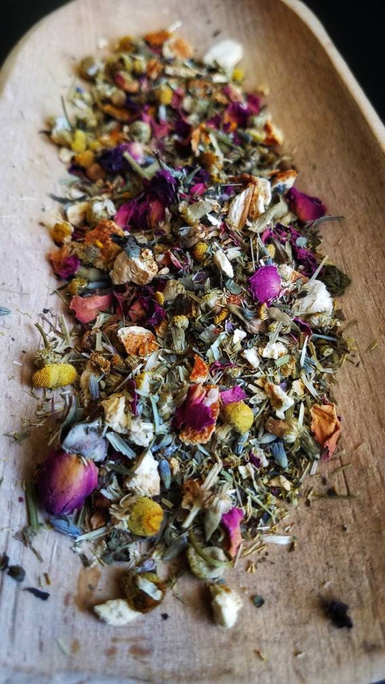 Op de afbeelding: Een houten lepel gevuld met een mengsel van gedroogde kruiden, bloemen en fruitschillen. Het mengsel bevat gedroogde rozenblaadjes, kamillebloemen, sinaasappelschil en andere kruiden.
