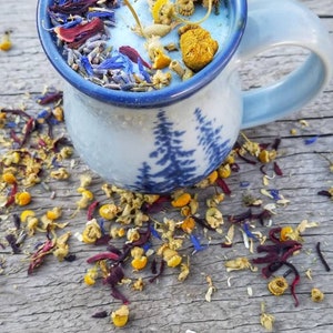 May include: A blue ceramic mug with a tree design filled with dried flowers and herbs. The mug is sitting on a wooden surface.