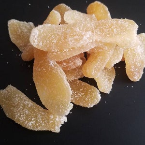 May include: A pile of candied ginger slices, coated in granulated sugar, against a dark background. The ginger pieces are a light yellow color, with a crystallized sugar coating. The image highlights the texture and sweetness of the snack.