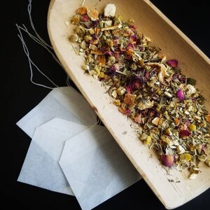May include: A wooden bowl filled with a blend of dried herbs, flowers, and citrus peel. The bowl is sitting on a black surface next to three empty tea bags.
