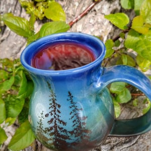 May include: A blue ceramic mug with a forest scene and a dark red liquid inside. The mug is sitting on a tree trunk with green leaves in the background.