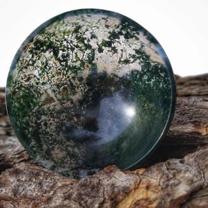 May include: A green moss agate sphere with a polished surface, displaying a variety of green and white patterns. The sphere is resting on a piece of rough, brown bark.