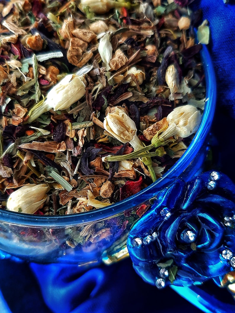 Pode incluir: Uma ch&aacute;vena de ch&aacute; de vidro azul cheia de uma mistura de ervas secas, flores e peda&ccedil;os de fruta. A ch&aacute;vena de ch&aacute; tem um design de rosa decorativo no lado.