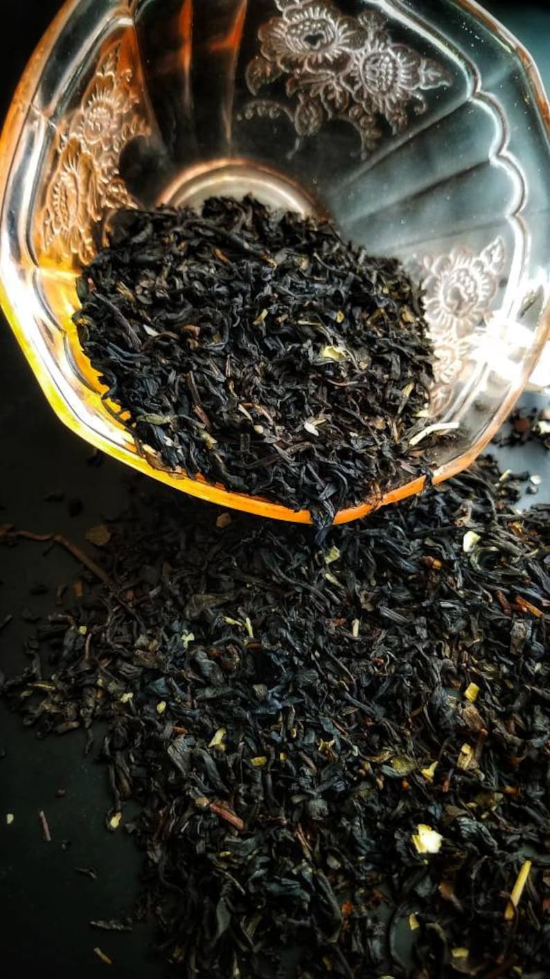 May include: Loose black tea leaves in a clear glass bowl with a floral design. The tea leaves are spilling out of the bowl onto a black surface.