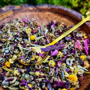 Pode incluir: Uma tigela de madeira cheia de uma mistura de ervas e flores secas, incluindo p&eacute;talos roxos, flores de camomila amarelas e folhas verdes. Uma colher de ouro est&aacute; descansando na tigela.