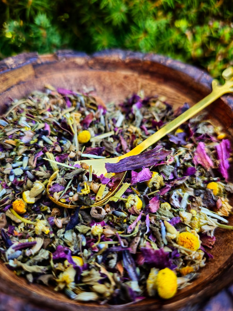 Pode incluir: Uma tigela de madeira cheia de uma mistura de ervas e flores secas, incluindo p&eacute;talos roxos, flores de camomila amarelas e folhas verdes. Uma colher de ouro est&aacute; descansando na tigela.