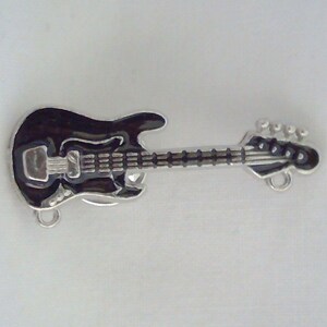 May include: A miniature black bass guitar charm with a silver border. The charm features a detailed fretboard and headstock, and two small loops for attachment. The guitar is set against a white background.