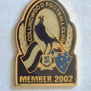 May include: A gold-coloured Collingwood Football Club member badge. The badge features a black and white magpie, the club's logo, and the text "COLLINGWOOD FOOTBALL CLUB" around the magpie. The badge also reads "MEMBER 2002".