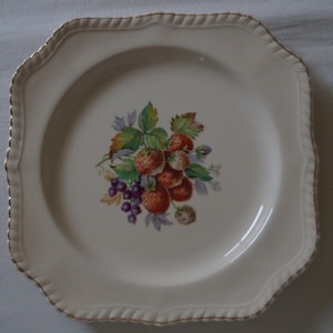 May include: A white square plate with a gold rim and a floral design featuring strawberries and blackberries.