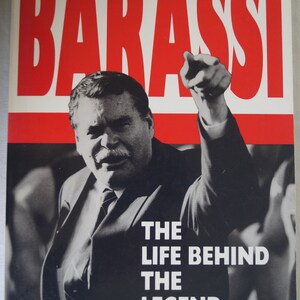 May include: A book cover with a red and white background. The title "BARASSI" is in large white letters on a red background. The subtitle "THE LIFE BEHIND THE LEGEND" is in black letters on a white background. The author's names "RON BARASSI & PETER McFARLINE" are in black letters on a white background.