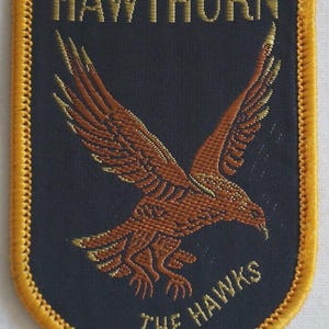 May include: A shield-shaped embroidered patch with a navy blue background and a gold border. It features a brown eagle with outstretched wings, and the words "HAWTHORN" and "THE HAWKS" in gold thread.