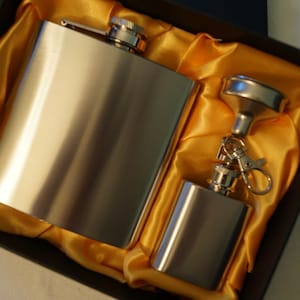 May include: Two silver metal hip flasks in a black box lined with gold satin. The larger flask has a cap and the smaller flask has a funnel and a key chain.