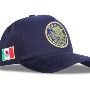 May include: Navy blue baseball cap featuring the Mexican coat of arms in gold and the word "MEXICO" in a circular design. The cap also has the Mexican flag embroidered on the side. A stylish accessory.