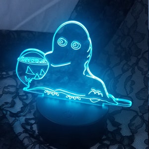 May include: A blue LED Halloween-themed night light featuring a ghost holding a jack-o'-lantern bucket. The ghost has large, circular eyes and a smiling mouth. The light sits on a round base.