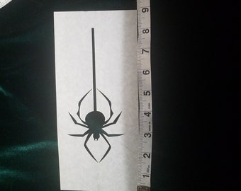 May include: A black spider silhouette on a white rectangular paper, approximately 4 inches (10 cm) in height, is measured with a tape measure. The spider design is centered and has a long vertical line extending from the top.