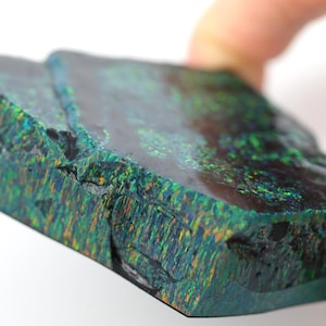 May include: A polished, rectangular gemstone with a dark, iridescent surface. The stone displays a play of color with flashes of green, blue, and orange. The edges show a rough, textured appearance. The gemstone is held by a hand.