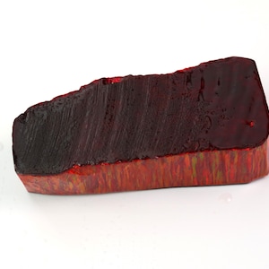 May include: A rectangular block with a dark, deep red top surface featuring visible brushstroke textures. The sides of the block display a vibrant, multi-colored pattern with streaks of red, orange, and yellow hues, creating a distinct contrast.
