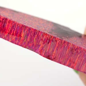 Könnte beinhalten: Ein rechteckiger Edelstein mit rauer Kante. Der Stein zeigt einen satten, rot-rosa Farbton mit orangefarbenen und violetten Streifen. Der Edelstein wird von einer Hand gehalten.