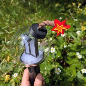 Könnte beinhalten: Ein klarer, kugelförmiger Leuchtstab mit schwarzem Griff und schwarzem Oberteil. Der Leuchtstab hat ein weißes und silbernes Logo im Inneren. Ein roter und gelber Stern ist oben befestigt, zusammen mit einem braunen, gedrehten Seil.