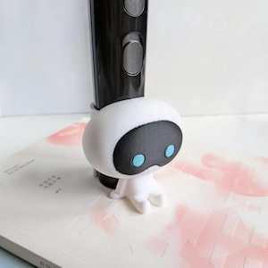 Könnte beinhalten: Eine weiße 3D-gedruckte Roboterfigur mit schwarzen Augen und blauen Pupillen. Der Roboter sitzt auf einer weißen Oberfläche mit rosa Flecken.