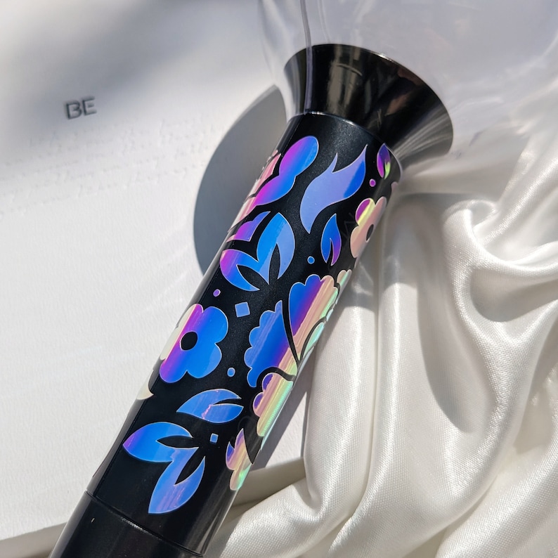May include: A black light stick with a floral design in iridescent colours. The light stick has a clear, rounded top and the text "BE" is visible in the background.