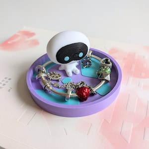 Può includere: Un porta anelli viola con un designo blu e bianco. Una piccola statuina bianca con occhi neri e un viso con un motivo bianco e nero si trova sul porta anelli. Un braccialetto d'argento con ciondoli è sul porta anelli. I ciondoli includono una fragola rossa, un pennello d'argento, una stella d'argento e un fiore d'argento.