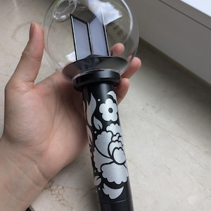 May include: A black and silver light stick with a clear globe top. The base features a floral design. The top has a black base with a logo. The light stick is held in a hand.