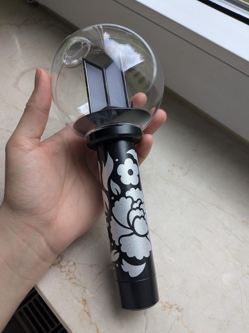 May include: A black and silver light stick with a clear globe top. The base features a floral design. The top has a black base with a logo. The light stick is held in a hand.
