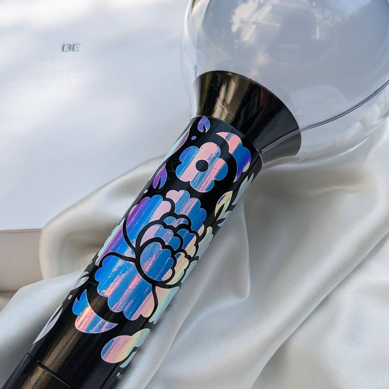 May include: A black light stick with a clear, spherical top and a floral design. The floral design features iridescent blue, pink, and purple colours. The light stick is resting on a white satin fabric.