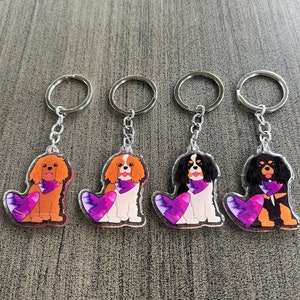 May include: Four keychains featuring Cavalier King Charles Spaniel dogs with purple hearts. Each keychain has a silver ring and a clear acrylic charm with a cartoon dog wearing a purple bandana.