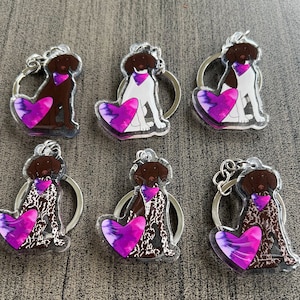 May include: Six keychains featuring a German Shorthaired Pointer dog breed with a purple and pink heart. The dogs are in different poses and have different colored collars. The keychains are made of clear acrylic.