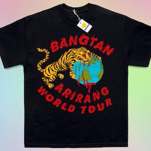 May include: Black t-shirt featuring a tiger with yellow and black stripes biting a globe. The globe has blue oceans and red blood dripping from it. Text in red reads "BANGTAN ARIRANG WORLD TOUR". A white tag hangs from the collar.