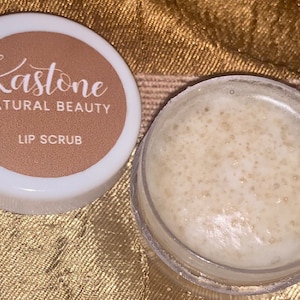 May include: A white jar with a brown lid. The lid has the text "Kastone Natural Beauty Lip Scrub" printed on it. The jar is filled with a white lip scrub.